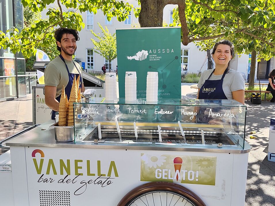Hinter einem Eiswagen mit dem Schriftzug Vanella Gelato stehen die Gelatieri von Vanella, links ein Mann, rechts eine Frau. Hinter ihnen steht mintgrünes ein Roll-Up mit dem AUSSDA Logo. Der Eiswagen befindet sich unter einem Baum auf dem Campus der Univ