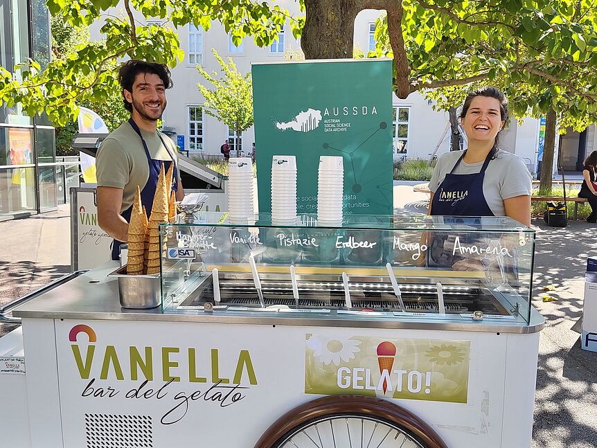 Hinter einem Eiswagen mit dem Schriftzug Vanella Gelato stehen die Gelatieri von Vanella, links ein Mann, rechts eine Frau. Hinter ihnen steht mintgrünes ein Roll-Up mit dem AUSSDA Logo. Der Eiswagen befindet sich unter einem Baum auf dem Campus der Univ