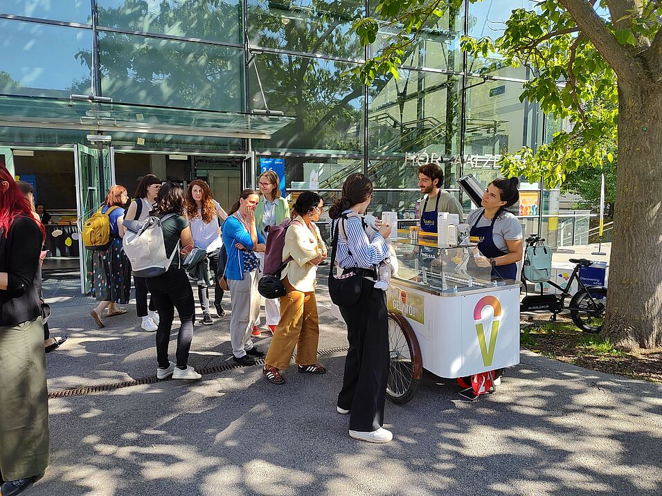 Eine Reihe von Menschen stehen vor dem Eiswagen von Vanella an. Die Gelatieri händigen Eis aus. Der Eiswagen befindet sich unter einem Baum auf dem Campus der Universität Wien.