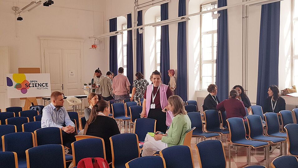 In der kleinen Aula auf dem Campus der Universität Wien sitzen und stehen Workshopteilnehmer*innen in Gruppen zusammen und diskutieren. Im Hintergrund ist das Logo des Open Science Festivals zu sehen.